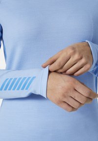 Camisa de manga larga de color azul claro con tejido texturizado. Presenta un puño con rayas azules y un diseño ajustado. Las manos están ajustando el puño.