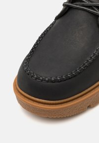 Czarne skórzane buty z zaokrąglonym czubkiem, z kontrastującą brązową gumową podeszwą i dekoracyjnym przeszyciem wzdłuż krawędzi.