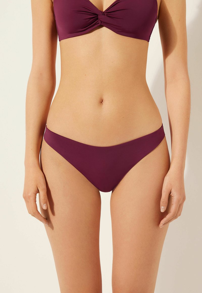 Le set de bikini mauve comprend un haut de bikini à devant torsadé et un bas taille basse assorti, fabriqué dans un tissu lisse et extensible offrant une couverture minimale.