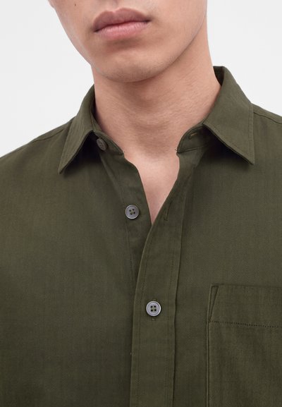 Gros plan d'une personne portant une chemise vert olive déboutonnée avec un col pointu et une poche poitrine.