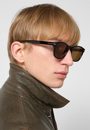 Giovane con capelli castano chiaro che indossa occhiali da sole scuri e una giacca di pelle marrone con il colletto alzato, mostrato di profilo.