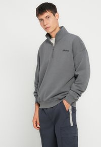 Sudadera gris con cremallera y cuello alto, con un logo sutil, ajuste relajado y puños acanalados. Combinada con pantalones cargo azul marino.
