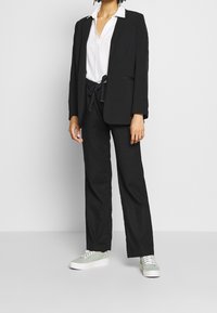 Blazer nero sopra una camicia bianca con bottoni, abbinato a pantaloni neri e sneakers verdi. Il blazer presenta una vestibilità su misura e una texture morbida.