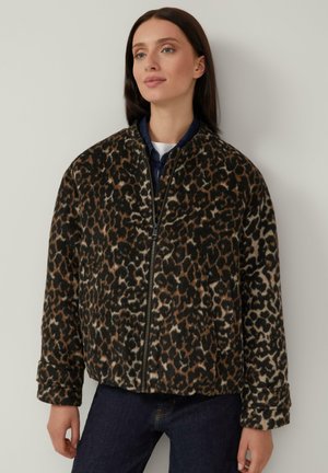 Vrouw in een leopardprint jack met rits en lange mouwen, in combinatie met donkere broek, staand tegen een effen lichte achtergrond.