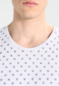 Hvid kortærmet skjorte med rund halsudskæring, der har et mønster af små blå bladmotiver, der er jævnt fordelt overstoffet.