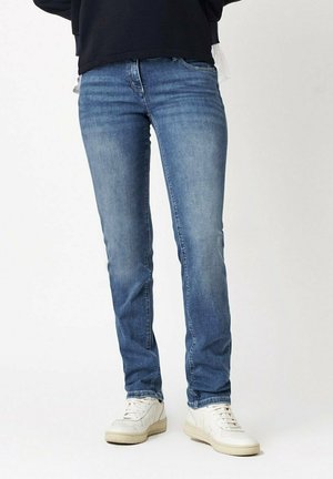 Persoon draagt strakke blauwe spijkerbroek en witte sneakers, staand tegen een effen lichte achtergrond.