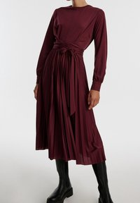 Burgundy pleated dress with long sleeves, a fitted bodice, and a waist tie. Styled with black knee-high boots for contrast.