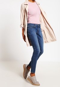 Osoba oblečená v světle béžovém trenčkotě, růžovém žebrovaném topu, modrých skinny džínách a béžových teniskách, stojící s jednou nohou mírně pokrčenou.