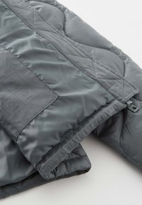 Grijze gewatteerde pufferjas met een gladde, glanzende textuur, voorzien van een klep met onderhoudsinstructies en een kleine haak aan de rand.