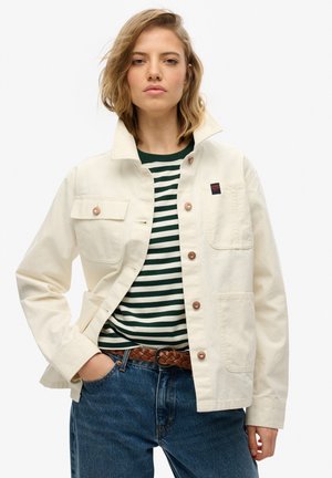 Beige Canvas-Jacke mit zwei Fronttaschen, bronzefarbenen Knöpfen, über einem grün-weißen gestreiften Hemd getragen, kombiniert mit blauen Jeans und einem geflochtenen Gürtel.