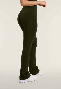 Mustat korkeavyötäröiset leggingit, joissa on sileä pinta ja liehuva leikkaus, yhdistettynä valkoisiin urheilukenkiin. Takana erottuva logoyksityiskohta.