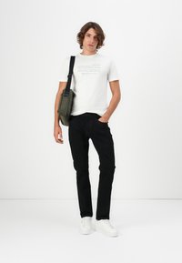 T-shirt blanche avec texte imprimé, jeans noirs et baskets blanches. Comprend également un sac à épaule olive avec une sangle.