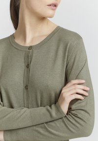 Olivově zelený kardigan s kulatým výstřihem, zapínáním na šest knoflíků vpředu a žebrovanými manžetami a lemem, vyrobený z měkkého, strukturovaného pleteného materiálu.