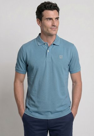 Hombre con una camiseta polo azul claro de manga corta con un pequeño emblema en el pecho y pantalones azul oscuro, de pie con las manos en los bolsillos.