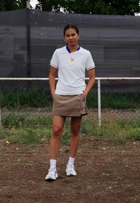 Biele tričko Nike s modrým golierom, s jemným logom a francúzskym emblémom, skombinované s hnedou sukňou a bielymi športovými topánkami.