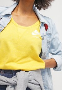 Geel Nike T-shirt met het "JUST DO IT" logo, gecombineerd met een lichtblauwe spijkerjack en een grijze trui die om de taille is gebonden.