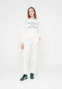 Sweatshirt de couleur crème avec un graphisme bleu "Converse All Star", accompagné d'un pantalon crème et de baskets vertes. Tissu lisse, coupe décontractée.