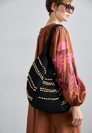 Sort vævet taske med træperler, der har en afslappet form og tekstureret design. Remmen er bred og komplementerer en brun kjole med blomstermønstre.