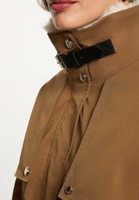 Giacca marrone con colletto alto, dotata di una fibbia nera, bottoni a pressione metallici e una finitura testurizzata. Accenti di pelliccia sono visibili.