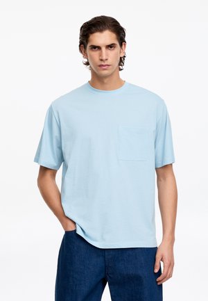 Jeune homme aux cheveux foncés portant un t-shirt bleu clair à manches courtes avec une poche poitrine et un pantalon bleu foncé, debout devant un fond blanc.
