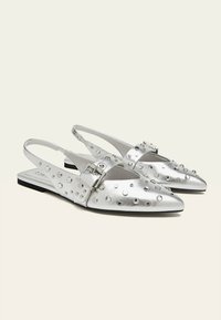 Ballerine slingback argento con punta affusolata, in pelle metallica testurizzata, con dettagli borchiati e una fibbia regolabile. Suola nera.