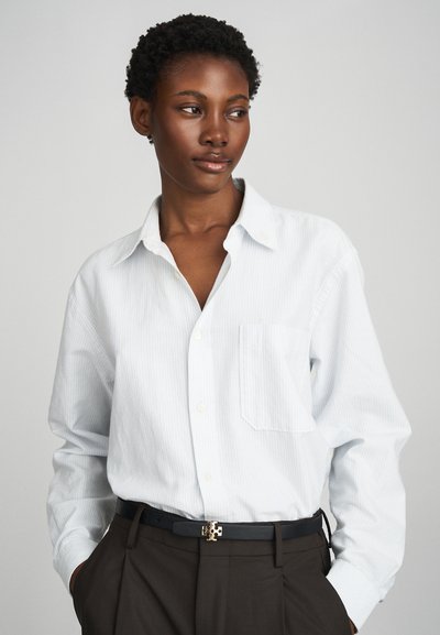 Bílá pruhovaná košile na knoflíky s kapsou na hrudi, spárovaná s tmavými kalhotami a černým opaskem se zlatou sponou.