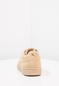 Sneaker Puma in suede beige vista dal retro, con un logo Puma dorato e una suola texturizzata, su uno sfondo bianco semplice.