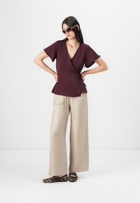 Burgundy wrap top with short sleeves, textured fabric; paired with wide-legged beige trousers and leopard print flats.