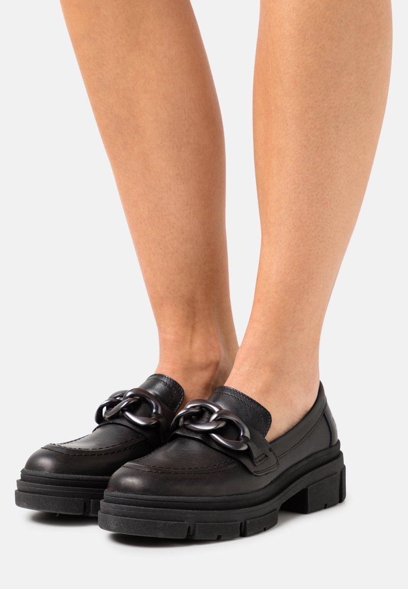 Mocassins en cuir noir avec semelles épaisses et détail de chaîne noire surdimensionnée, portés pieds nus sur un fond clair.