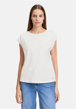 Weißes Baumwoll-T-Shirt mit kurzen Kappärmeln und einem runden Ausschnitt, das auf der linken Seite ein strukturiertes Blumenmuster aufweist. Darunter werden blaue Jeans getragen.