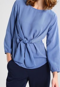 Žena v modro-bílé pruhované blůze s uzlem vpředu a dlouhými rukávy, kterou doplnila tmavě námořnickými kalhotami.