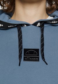 Un sweat à capuche bleu avec une surface texturée, des cordons noirs avec des accents blancs et un patch en caoutchouc noir sur le devant.