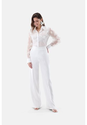 Frau trägt eine durchsichtige weiße Bluse mit Blumenstickerei, hoch taillierte weiße Hose, große Blumenohrringe und weiße offene Absätze, lächelnd und posierend.