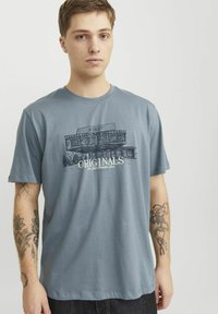 Tricou din bumbac albastru, cu un imprimeu grafic al unei clădiri și cuvântul "ORIGINALS" în alb. Mâneci scurte și croială lejeră.