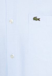 Camisa de botones azul claro con una tela texturizada, que presenta un pequeño logo de cocodrilo verde en el bolsillo del pecho. Se pueden ver cuatro botones al frente.