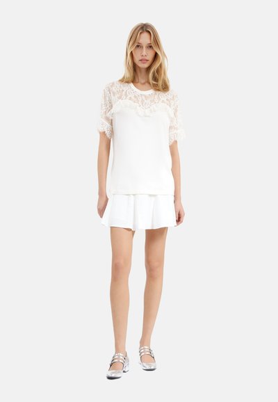 Hvid t-shirt med blonde korte ærmer, flæset halsudskæring, parret med en hvid nederdel. Modellen har sølvfarvede flade sko på og har bølget hår.