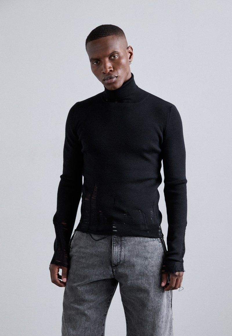 Μαύρο πλεκτό πουλόβερ με ψηλό λαιμό και φθαρμένο στρίφωμα και λεπτομέρειες στα μανίκια. Συνδυάζεται με ανοιχτό γκρι, relaxed-fit τζιν παντελόνι.