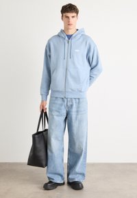 Lichtblauwe rits hoodie met een kap, voorkzakken, gecombineerd met losse lichte spijkerbroek en zwarte instappers, terwijl een zwarte totebag wordt vastgehouden.