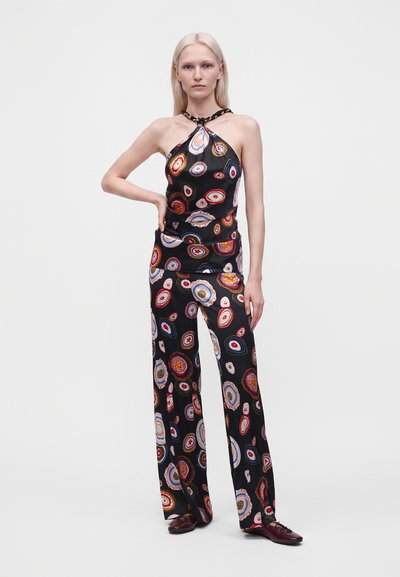 Femme portant un haut dos nu noir et un pantalon à jambes larges avec des motifs circulaires colorés, debout avec une main sur la hanche sur fond blanc.