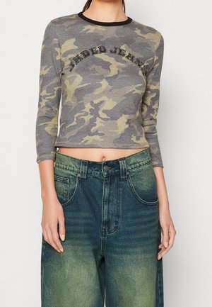 Femme portant un crop top manches longues camouflage gris et vert avec le texte "Jaded Jeans" et un jean bleu délavé taille haute.