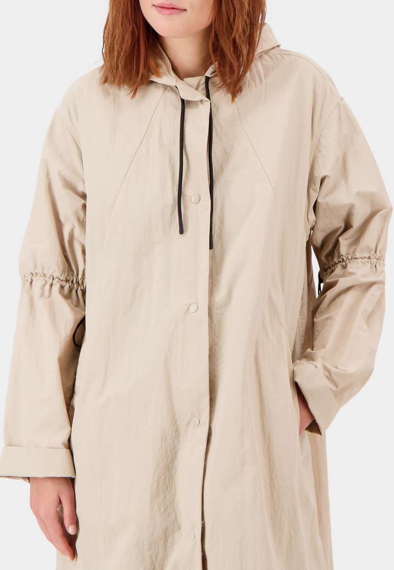 Beige leichter Trenchcoat mit Kapuze, Knopfverschluss und gerafften Details an den Ärmeln. Hergestellt aus zerknittertem Stoff mit lockerer Passform.