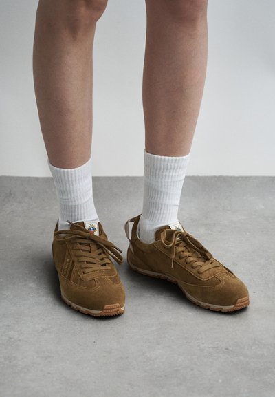 Ténis de camurça castanho com um design de atacadores, apresentando um logo branco e sola de borracha texturizada, combinados com meias brancas caneladas.