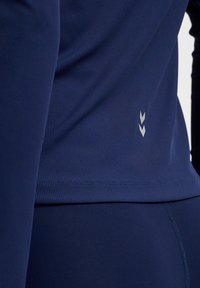 Close-up van een persoon die een nauwsluitende marineblauwe sporttop en legging draagt met een klein reflecterend dubbel-chevronlogo op de rug.