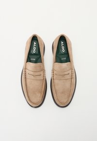 Mocassins en suède beige à enfiler, avec un bout arrondi, des détails cousus et des semelles en caoutchouc noires contrastantes. Semelles intérieures de marque vertes visibles.