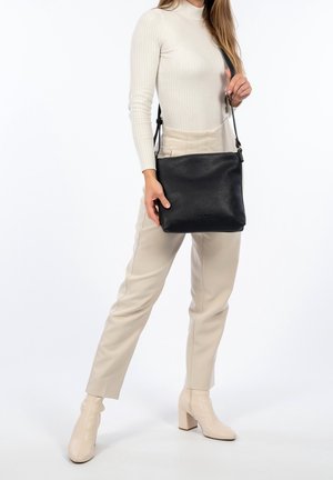 Frau in beigen Hosen und Stiefeln hält schwarze Leder-Umhängetasche mit verstellbarem Riemen vor weißem Hintergrund.