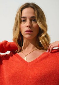 Roter V-Ausschnitt-Pullover aus weichem Material mit einer strukturierten Oberfläche. Eine goldene Anhänger-Halskette sorgt für einen Kontrast zu der lebhaften Farbe.