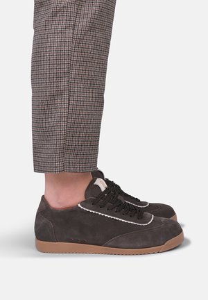 Persona che indossa pantaloni marroni a quadri e sneakers in camoscio marrone scuro con lacci neri e suole in gomma marrone chiaro, in piedi su uno sfondo chiaro.