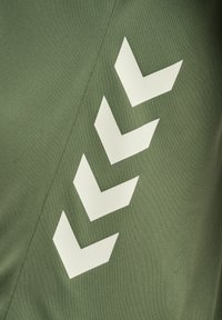 Olive-greenowy materiał z teksturowaną powierzchnią i dużymi, białymi wzorami w kształcie ząbków po bokach, tworzącymi odważny grafiki design.