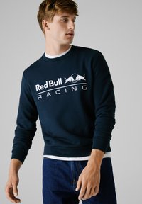 Sudadera azul marino con el logo blanco de "Red Bull Racing", cuello redondo y mangas largas, combinada con unos vaqueros oscuros.