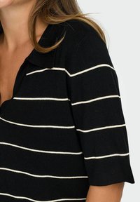 Woman wearing a black knit top with white horizontal stripes and a collar, showing shoulder and partial neckline.